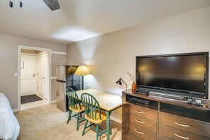 Habitación de hotel con cama, escritorio con sillas, televisor, lámpara y minibar.