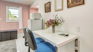 Ein modernes Zimmer mit weißem Schreibtisch, blauem Stuhl und einer Vase mit Blumen. Ein Fenster bietet Blick auf Bäume, eine weiße Klimaanlage und ein graues Schränkchen sind ebenfalls vorhanden.