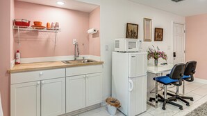 Eine Küche mit weißen Schränken, einem Spülbecken, einer Mikrowelle und einem Kühlschrank. Zwei Stühle und ein Mülleimer sind ebenfalls vorhanden.