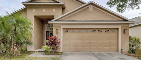 A residential house with a garage, a front entrance, and a small garden with palm trees and colorful plants.