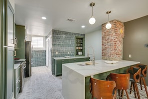 A kitchen with green cabinets, a brick accent wall, a white island with bar stools, and a window with blinds. - Fire Pit & Pergola! Renovated Brick Beauty By TTU (Lubbock)