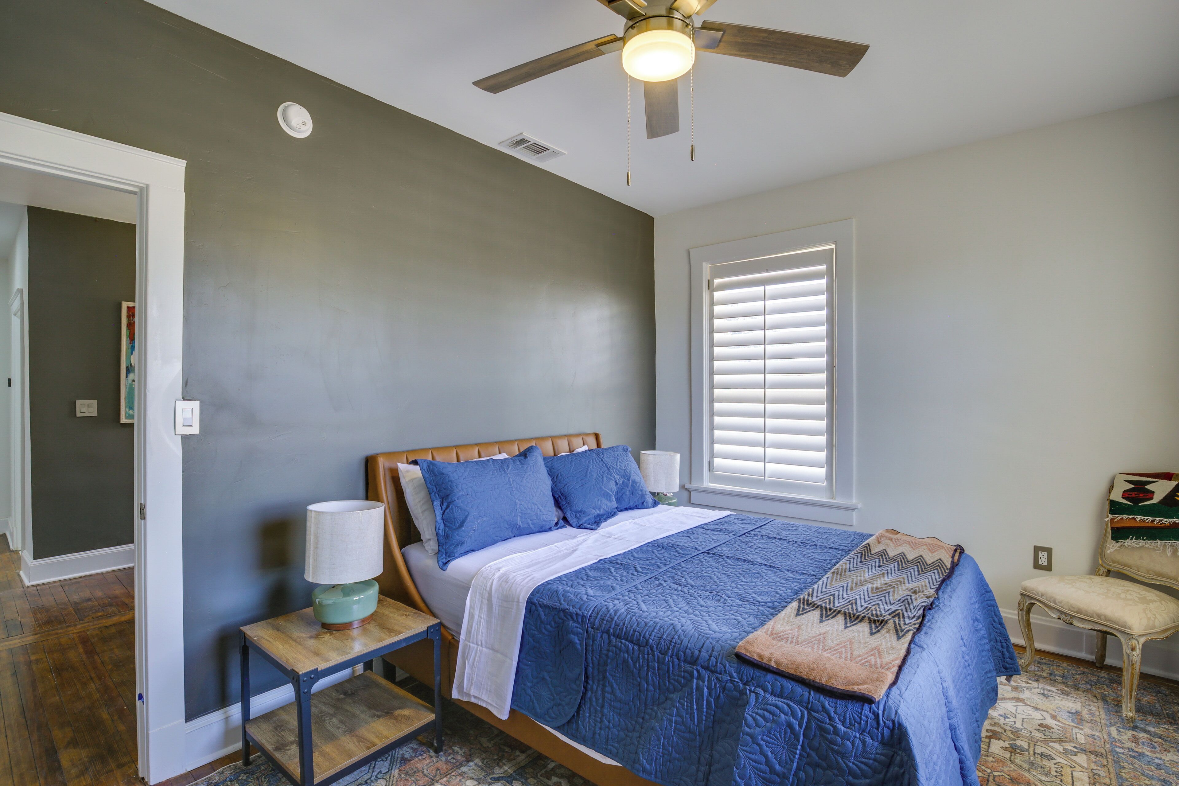 A bedroom with a bed, a nightstand, a chair, a ceiling fan, and a window with blinds.