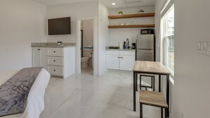 Quarto de hotel moderno com cama, escrivaninha, cozinha compacta e televisão na parede.