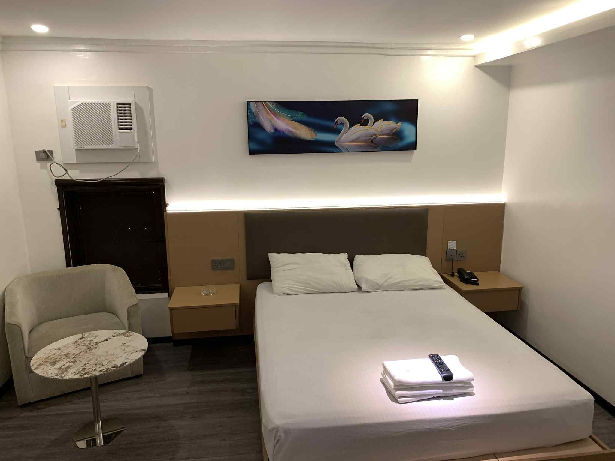 Una habitación de hotel con cama, una silla, una mesita, un cuadro y un equipo de aire acondicionado.