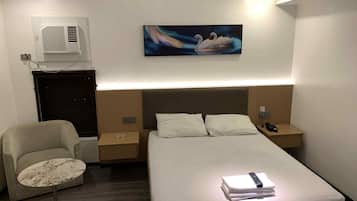 Una habitación de hotel con cama, una silla, una mesita, un cuadro y un equipo de aire acondicionado.