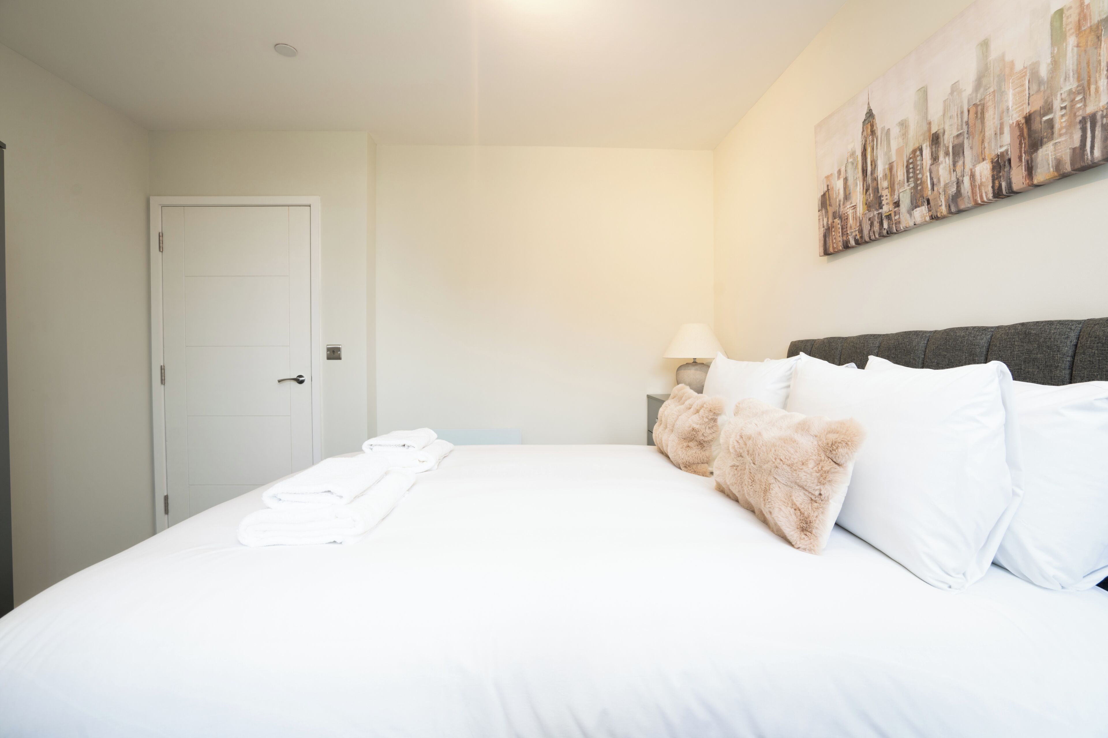 Een hotelkamer met een bed, witte lakens, een stadslandschap schilderij, een deur en een nachtkastje met een lamp.