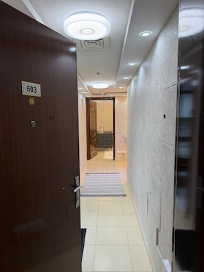 A hotel corridor with a wooden door labeled '603,' a tiled floor, a rug, a bathroom with a bathtub, and a ceiling with recessed lighting.