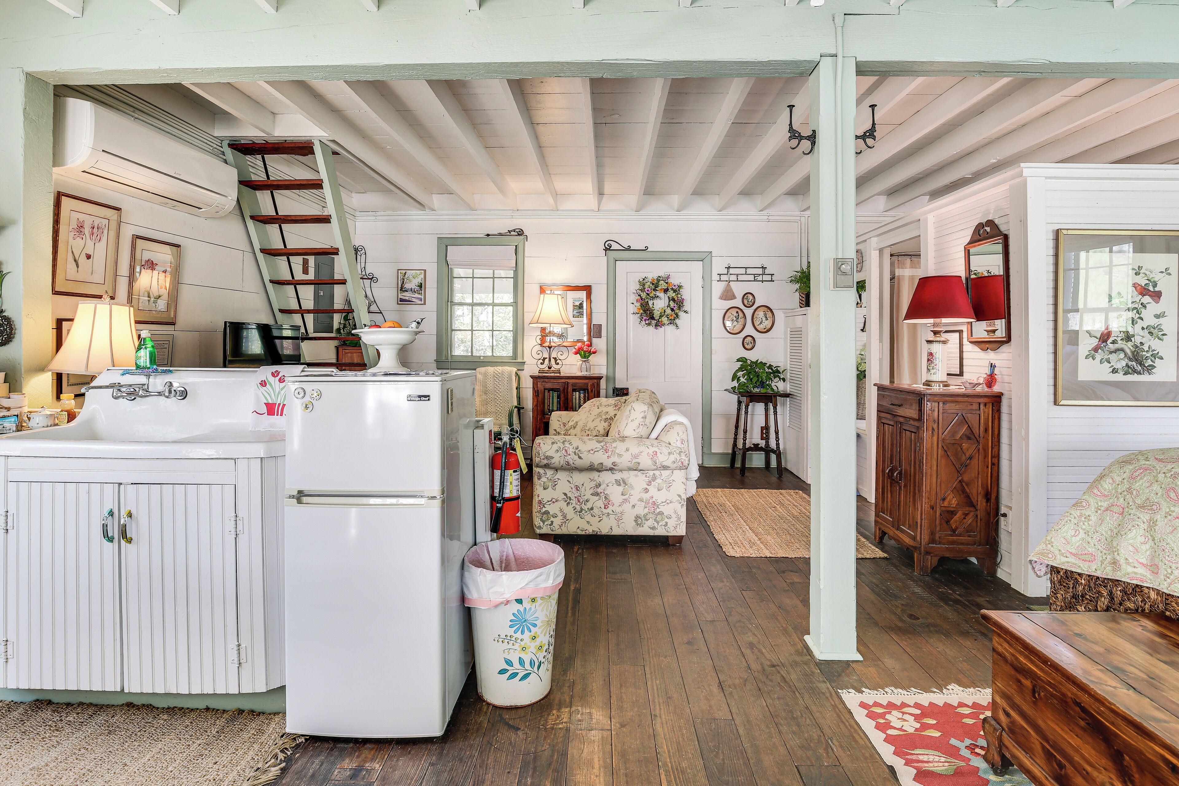 A kitchen with a refrigerator, stove, and sink; a wooden ladder; a floral-patterned sofa; a wooden cabinet; and a red lamp.
