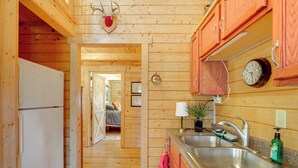Interior de una cabaña de madera con zona de cocina, que incluye un refrigerador, un lavamanos y un reloj.