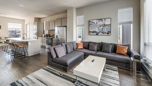 A modern kitchen with a central island, a dining area with bar stools, a gray sectional sofa with a coffee table, and a large abstract painting on the wall. - Work & Play! Gem w/ Fire Pit, 3 Mi to Dtwn Dallas (Dallas)