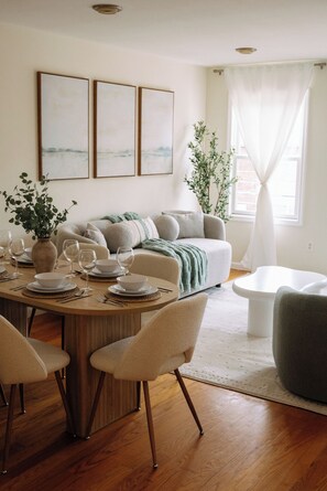 A dining area with a round table set for four, a sofa, and a window with sheer curtains. - Sleeps 20 I Metlife 10mins 6BR/4BA I 2 Free Parking & Private Deck (Jersey City)