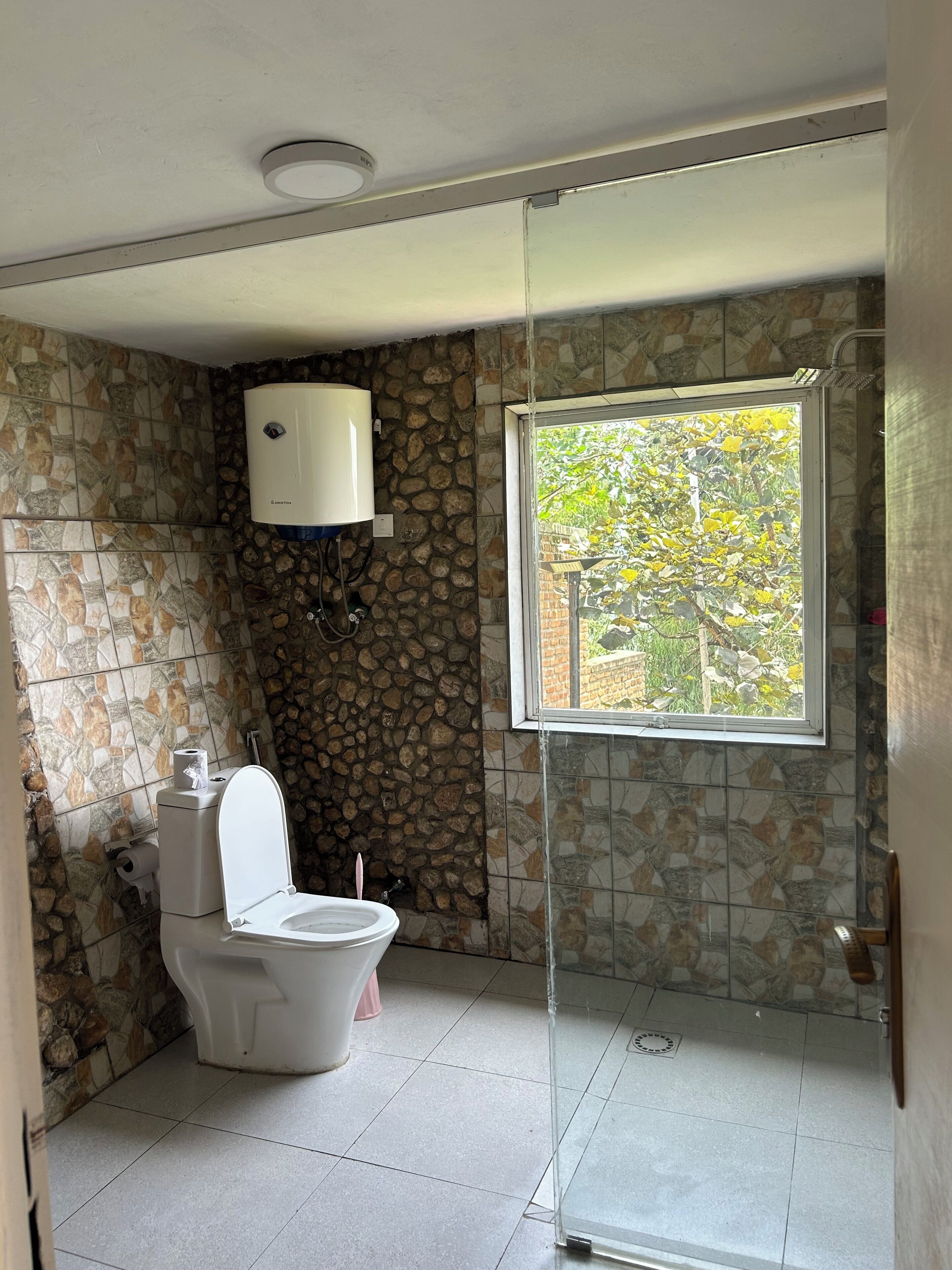 A bathroom with a toilet, a shower, a window with a view, and a stone wall.