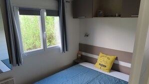 Un llit individual amb un edredó blau, un coixí groc amb text, una finestra amb cortines i una petita estant sobre el llit.