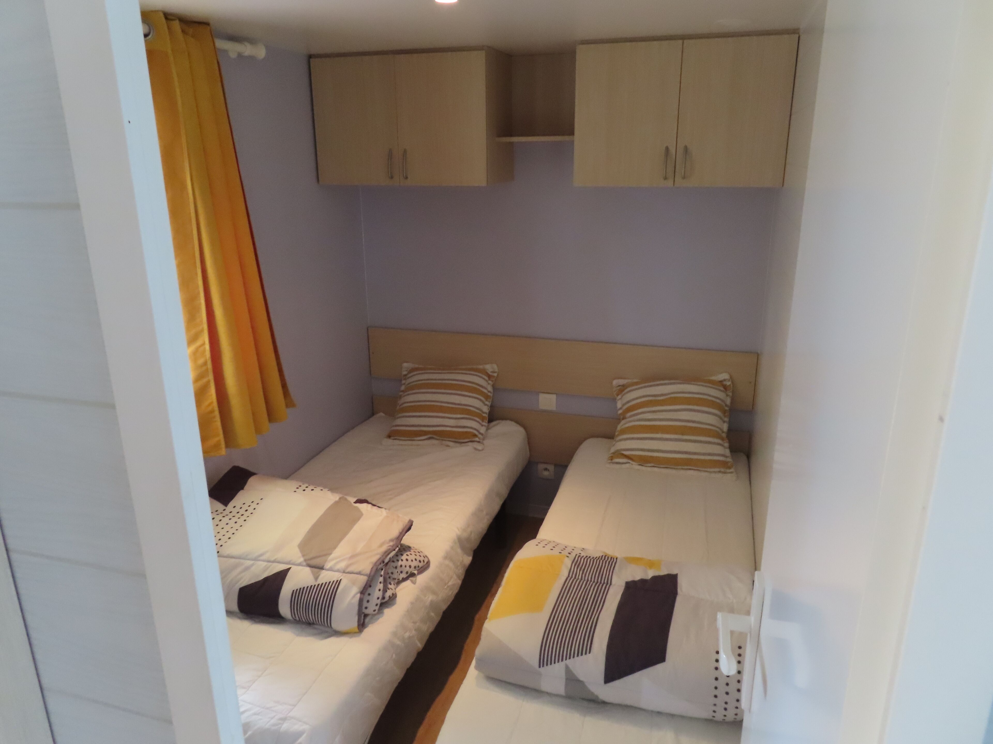 Ein kleines Zimmer mit zwei Betten, einem hölzernen Kopfteil und einem gelben Vorhang.