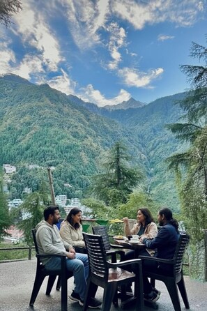 A group of people seated at an outdoor table with a mountainous landscape in the background. - Thrillomania Mcleodganj (Dharamshala)