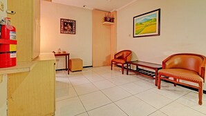 Ein Zimmer mit einem Holztisch, zwei Ledersesseln, einem kleinen Tischchen, einem Regal mit Dekorationsobjekten, einem gerahmten Gemälde und einem Feuerlöscher.
