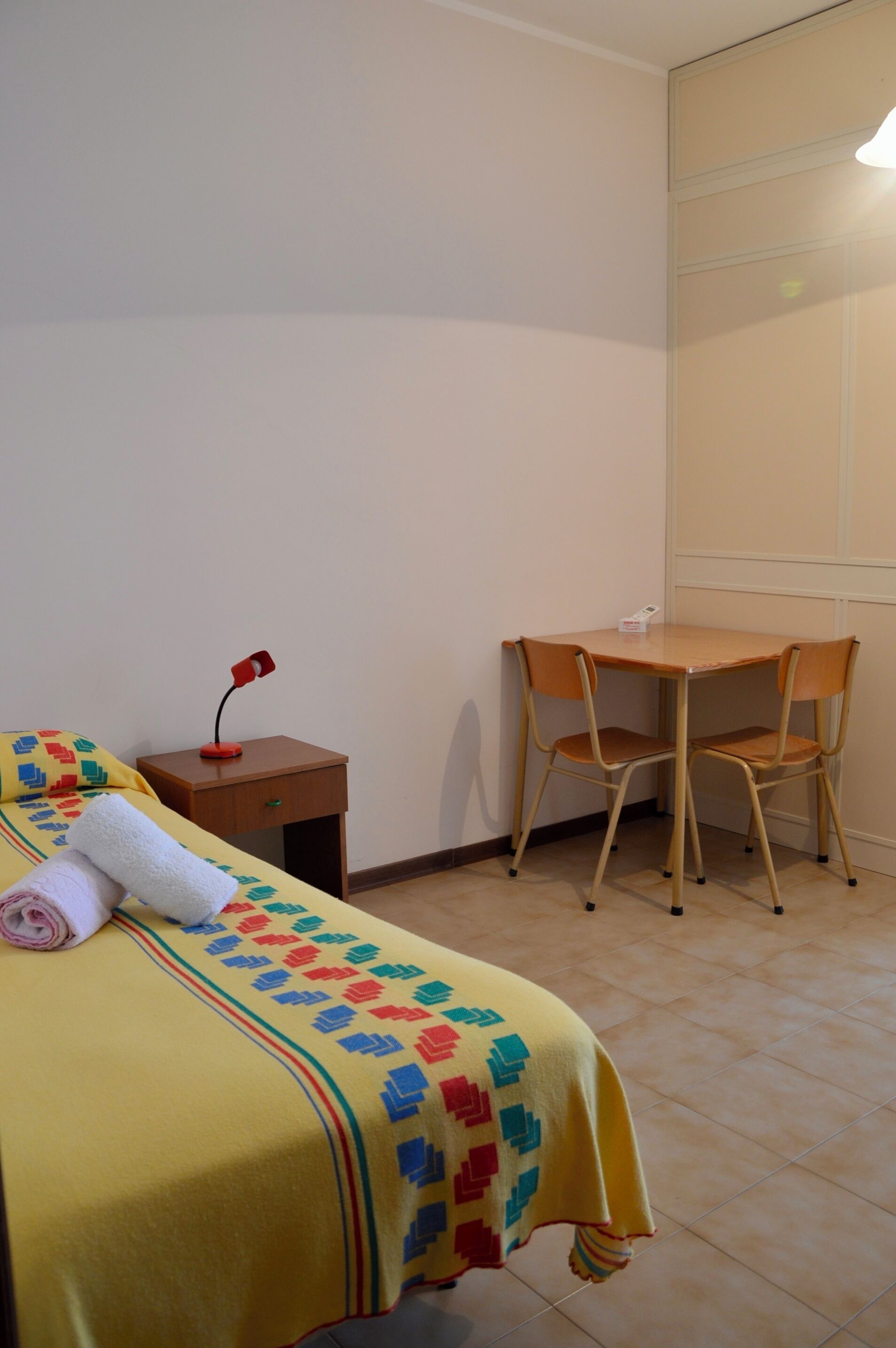 A room with a bed, a small wooden table, two chairs, a nightstand, and a wall-mounted shelf.