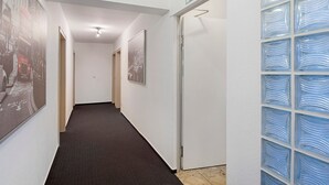 Un pasillo con piso alfombrado, cuadros en las paredes, una puerta con letrero de salida verde y una pared de bloques de vidrio a la derecha.