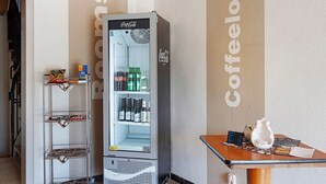 Una máquina expendedora con el logotipo de Coca-Cola, un refrigerador con estantes, y una mesa con una tetera y un menú.