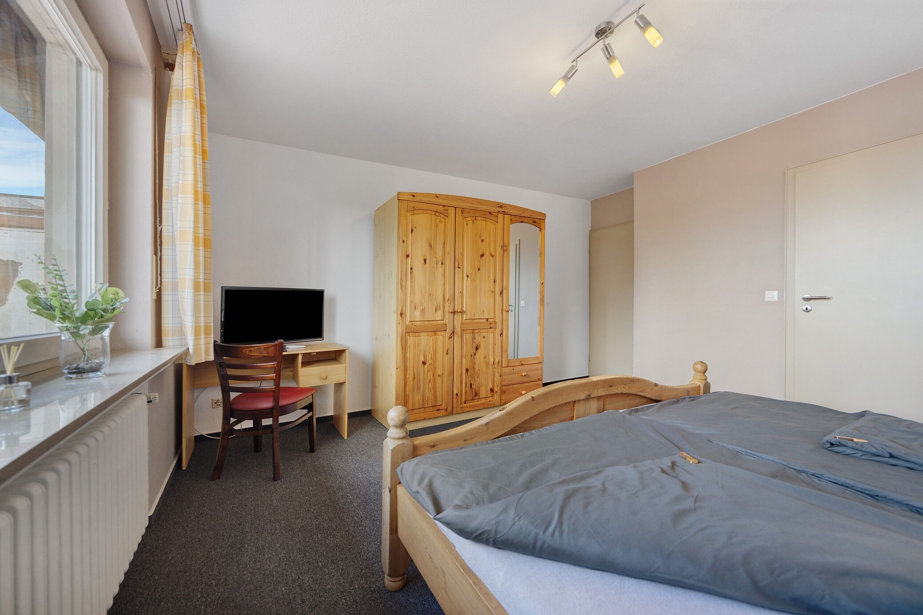 A bedroom with a bed, a wooden wardrobe, a desk with a chair, a television, and a window with curtains.