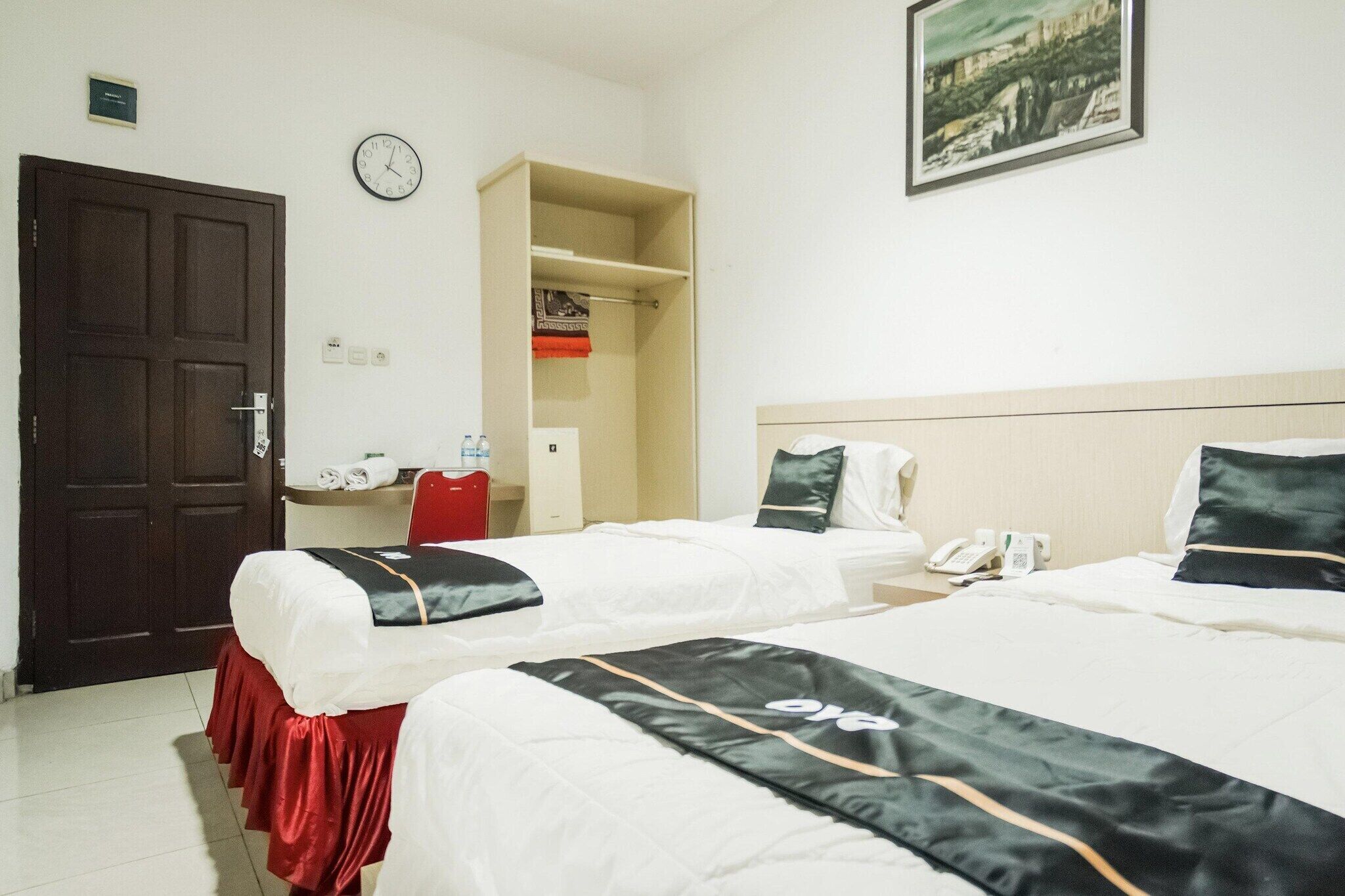 A hotel room with two beds, a wardrobe, a desk with a chair, a clock, and a framed picture on the wall.
