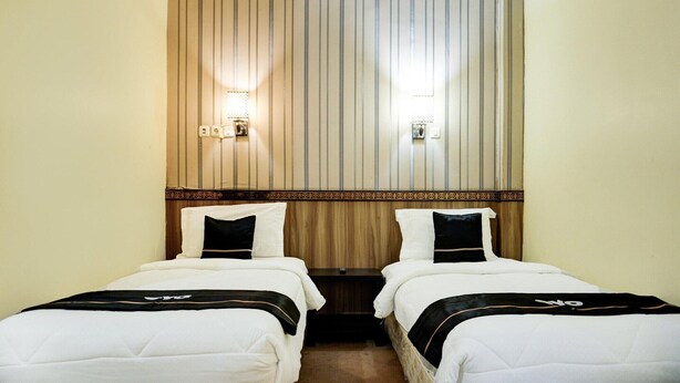 A hotel room with two single beds, wooden headboard, wall-mounted lights, and a small bedside table.