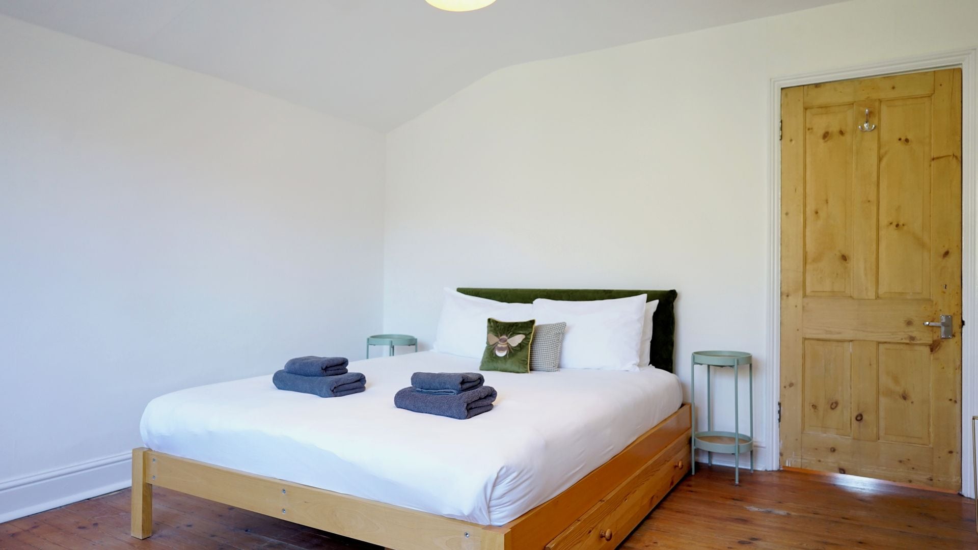 A single bed with a wooden frame, white bedding, and two folded towels. There is a green upholstered headboard, a wooden door, and two small green nightstands with lamps.