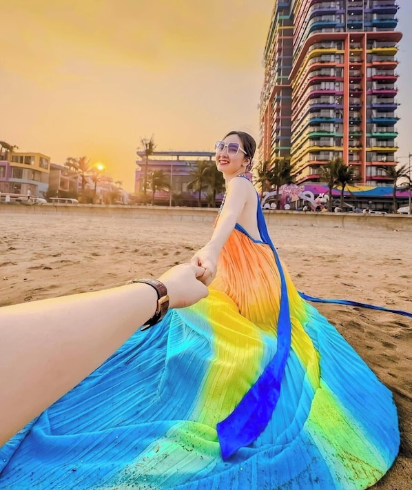 A person holding hands with another, wearing a colorful dress, on a sandy beach with buildings in the background.