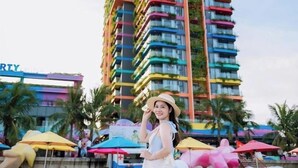 Un hotel de gran altura con varios balcones, una playa arenosa al frente y sombrillas de playa.