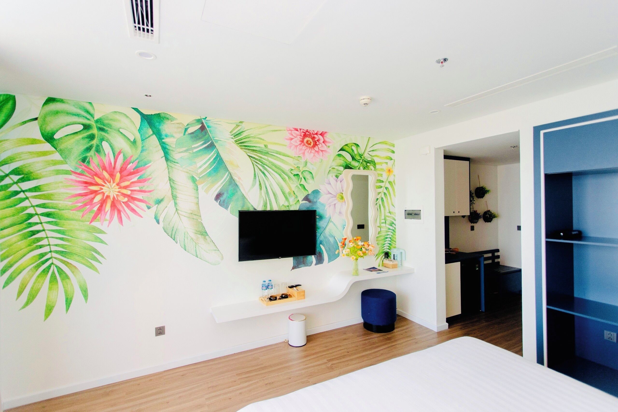 A modern hotel room with a large bed, a flat-screen TV mounted on the wall, a small kitchen area, and a bathroom visible through an open door.