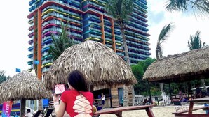 Una playa con sombrillas de paja, una mujer tomando una fotografía y un hotel colorido al fondo.
