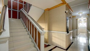 Eine Treppe mit weißen Stufen und einem hölzernen Geländer, die zu einer Zwischenebene mit einer beigen Wand und einer verzierten Decke führt.