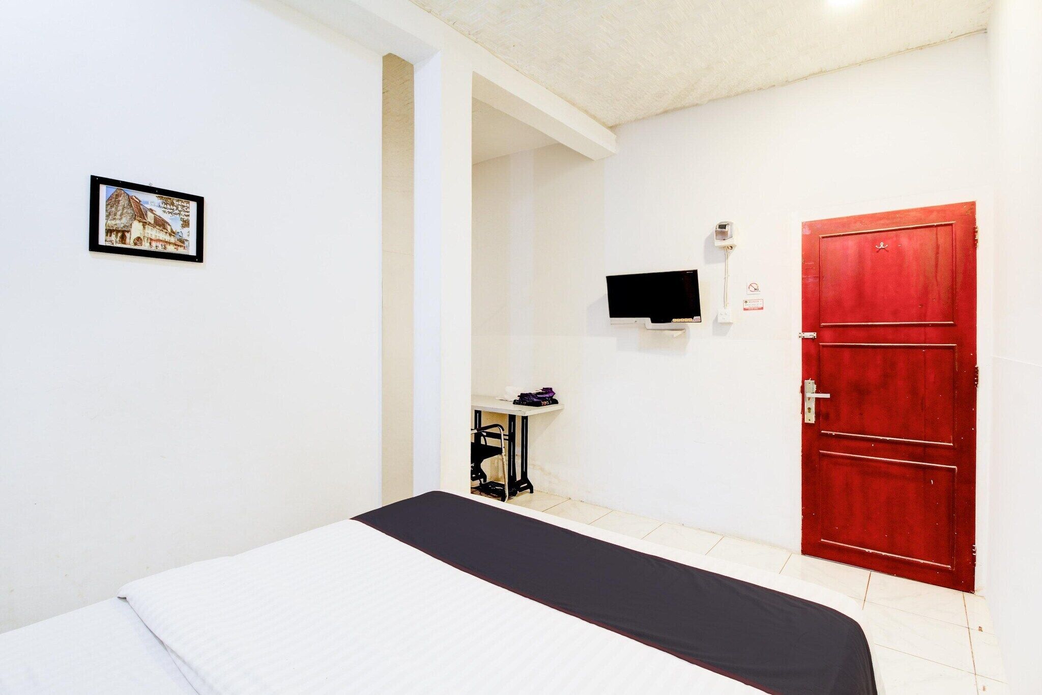 A room with a bed, a red door, a television, and a small table with a sewing machine.