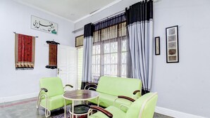 Quarto com sofá verde, mesinha redonda, tapete estampado e janela com cortinas.
