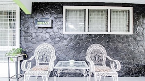 Mural de pedra com uma placa a indicar 'Guesthouse NELVI' e duas cadeiras de metal branco com uma mesa de vidro em frente.