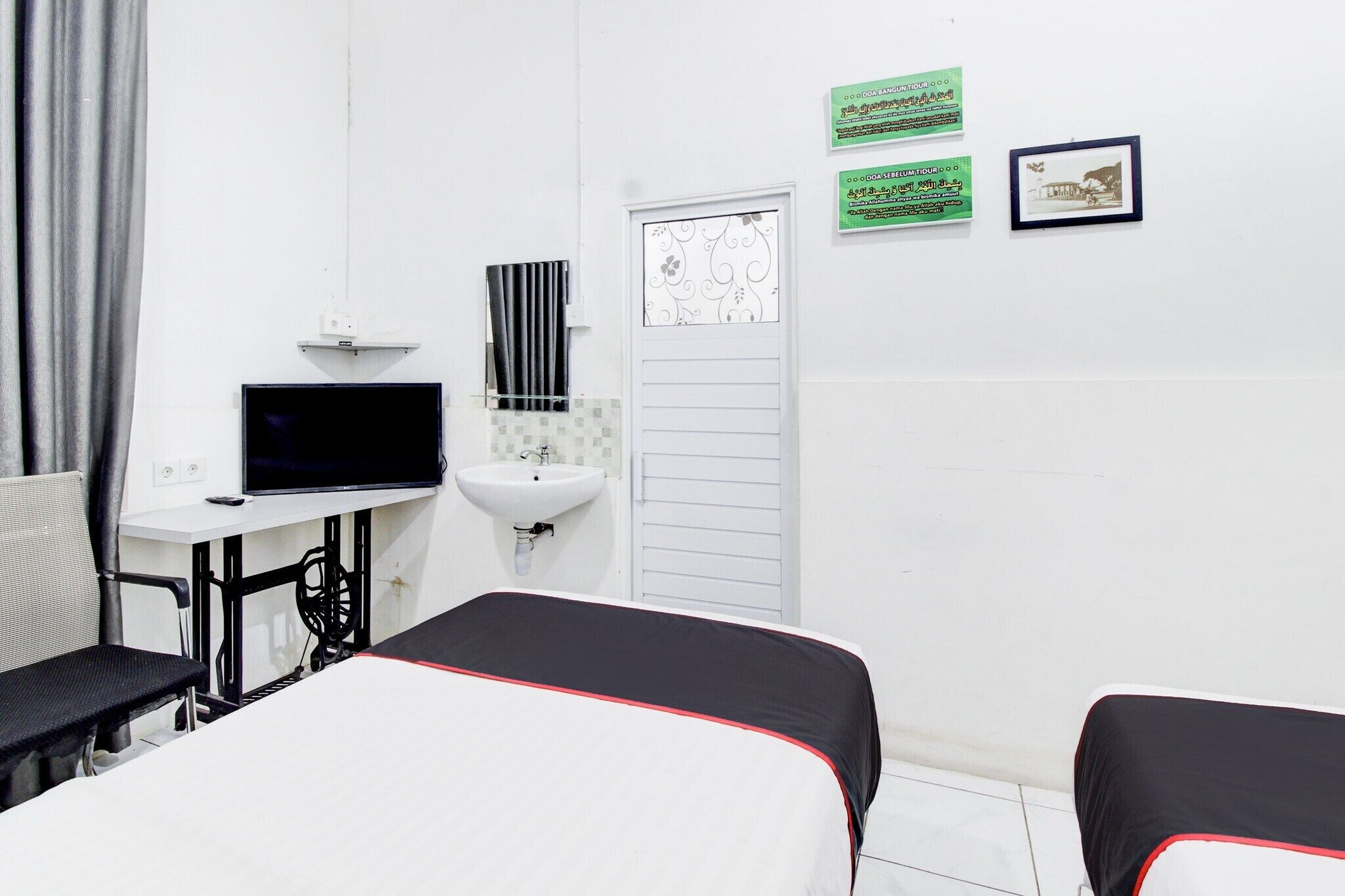 Ein Hotelzimmer mit Bett, Schreibtisch inklusive Computer, Waschbecken und zwei gerahmten Bildern an der Wand.