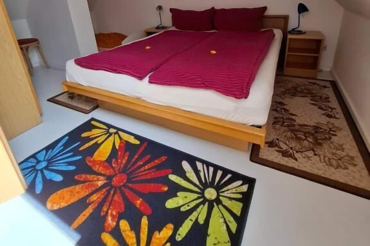 A bedroom with a sloped ceiling, a bed with a red blanket, two bedside tables with lamps, and a colorful floral rug.