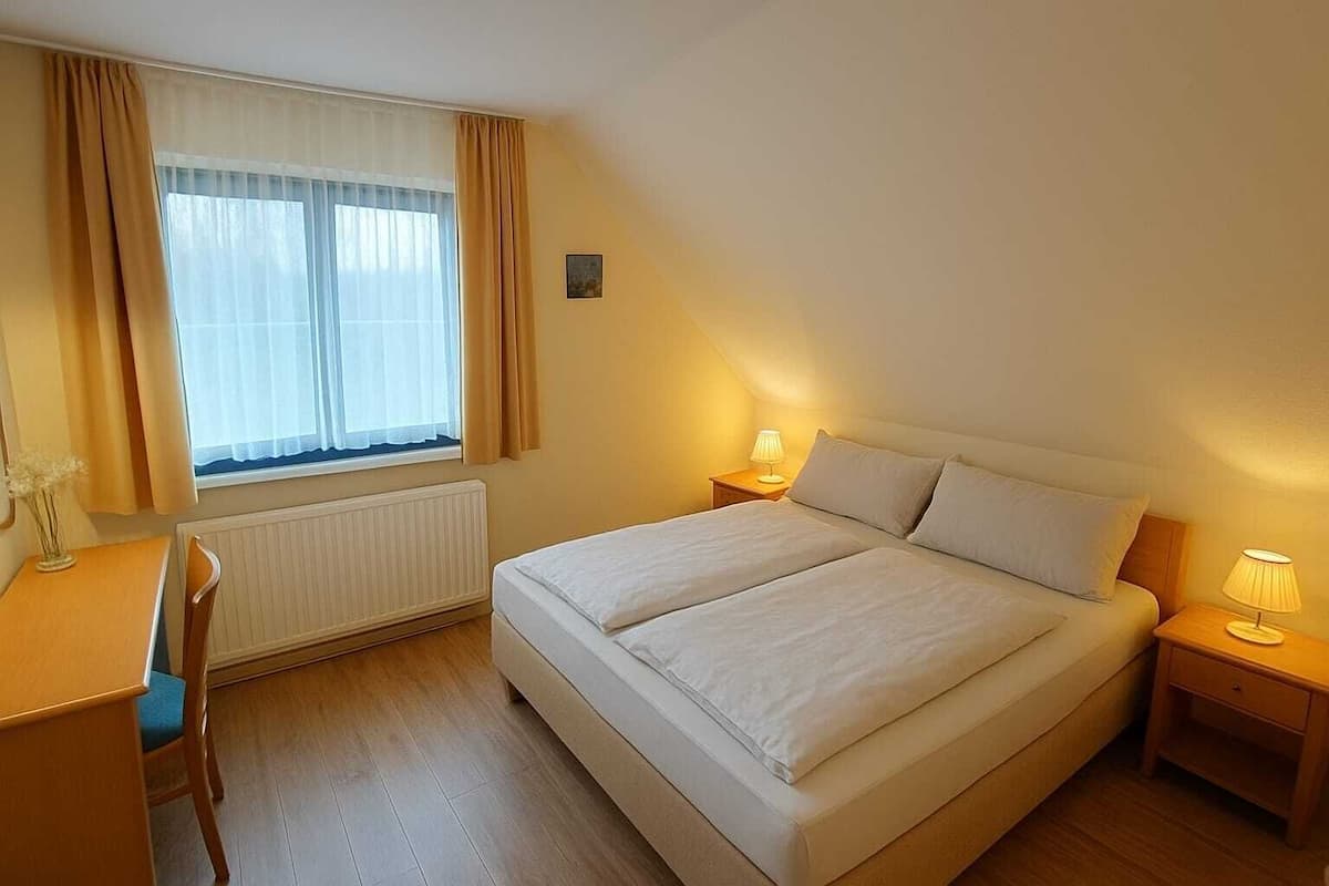 A bedroom with a bed, bedside lamps, a chair, a desk, a window with curtains, and a radiator.