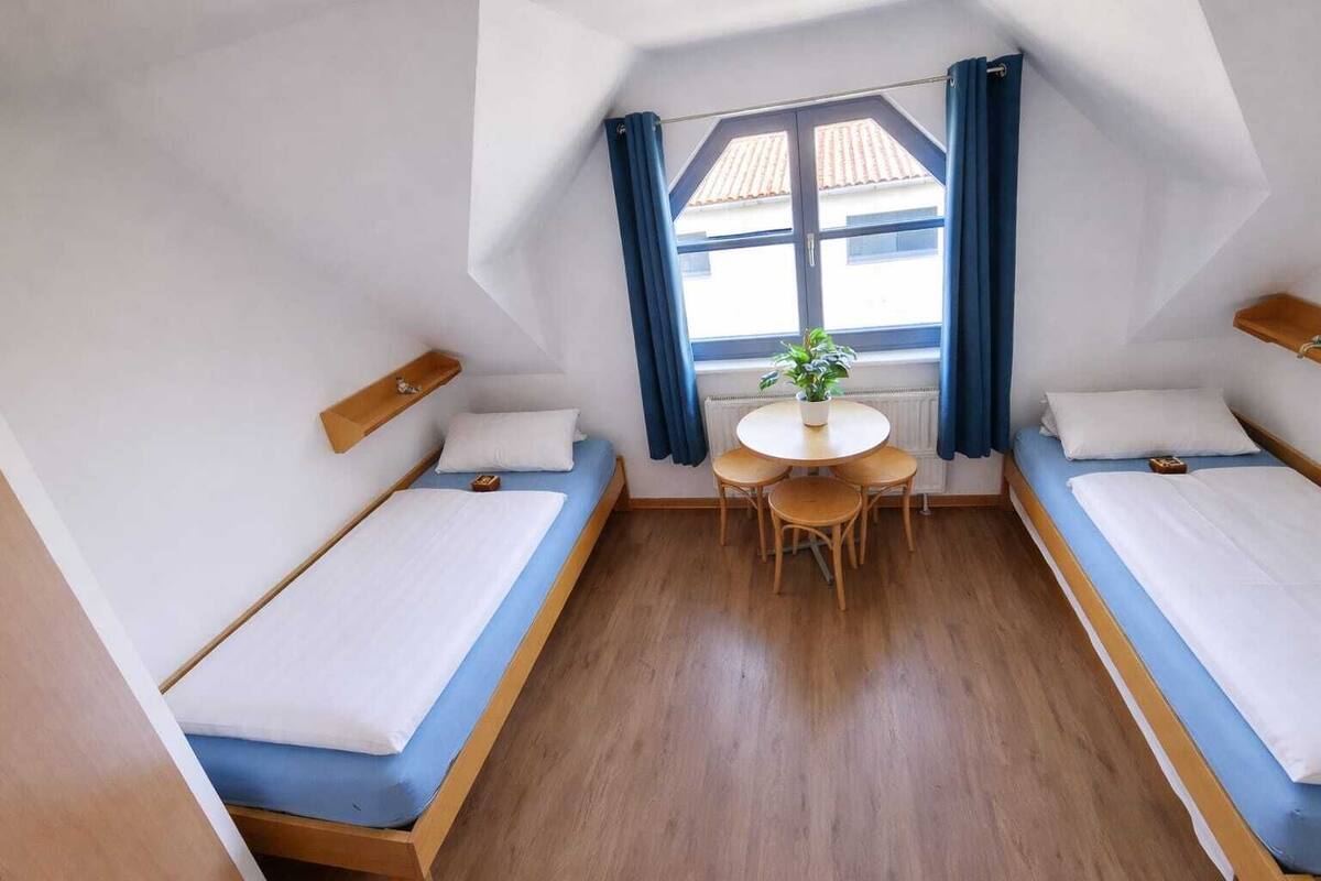 A room with two beds, a small table with chairs, a potted plant, and a window with curtains.
