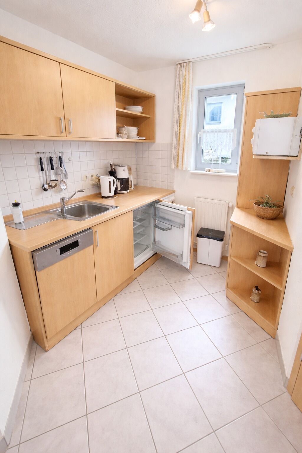 A compact kitchen with wooden cabinets, a dishwasher, a kettle, and a window with curtains.