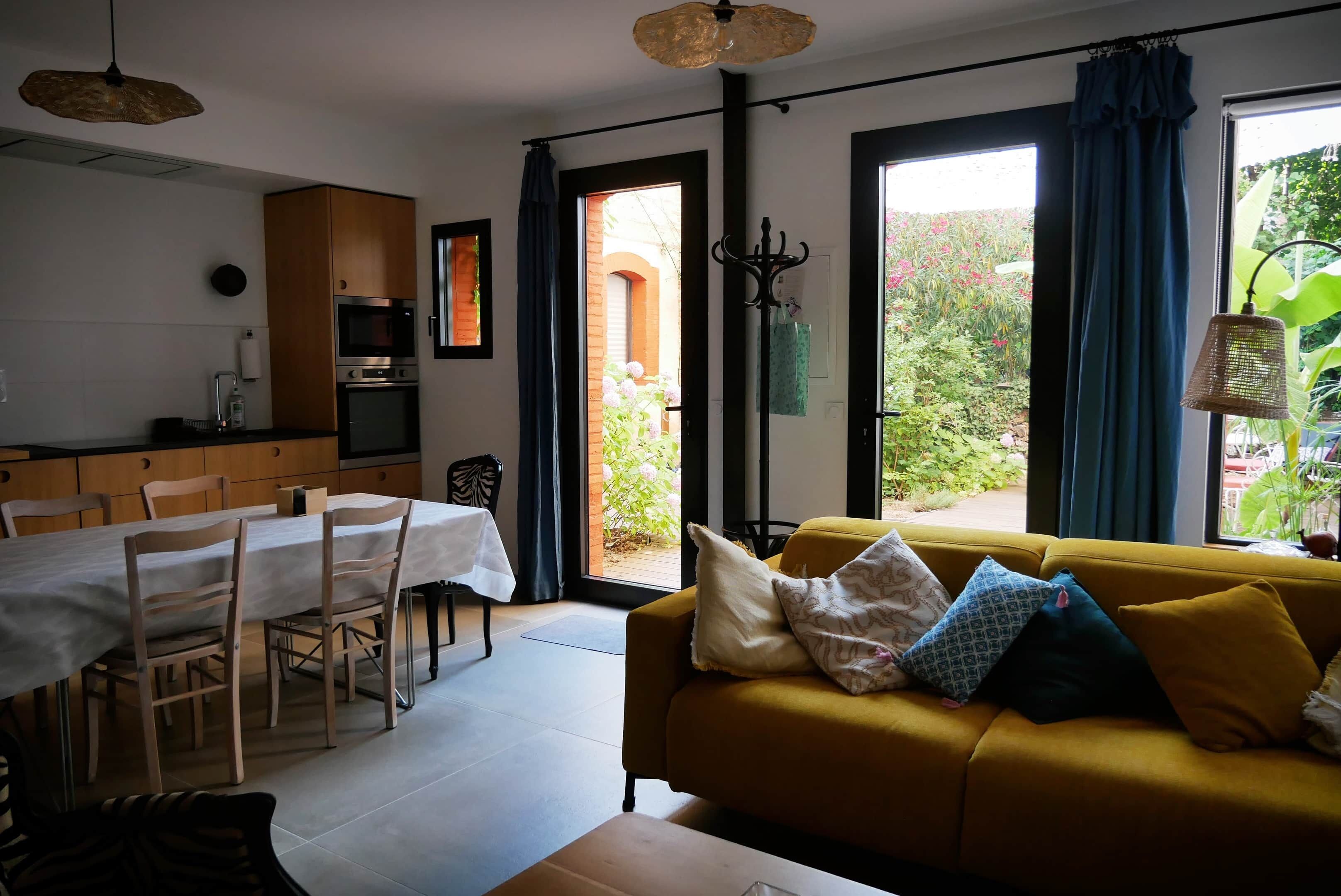 A dining area with a table and chairs, a sofa with cushions, a hanging light fixture, and a view through a doorway to an outdoor garden.