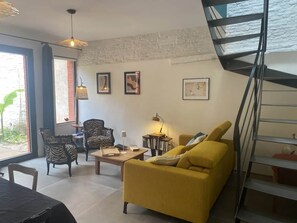 Una sala de estar con un sofá amarillo, dos sillones, una mesa de centro y una estantería. Se ve una escalera a la derecha y una ventana con vista a follaje.