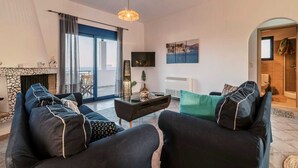 Ein Wohnzimmer mit einer Sofagarnitur, einem Fernseher, einem Kamin und Blick ins Freie.