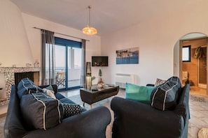 Ein Wohnzimmer mit einer Sofagarnitur, einem Fernseher, einem Kamin und Blick ins Freie.