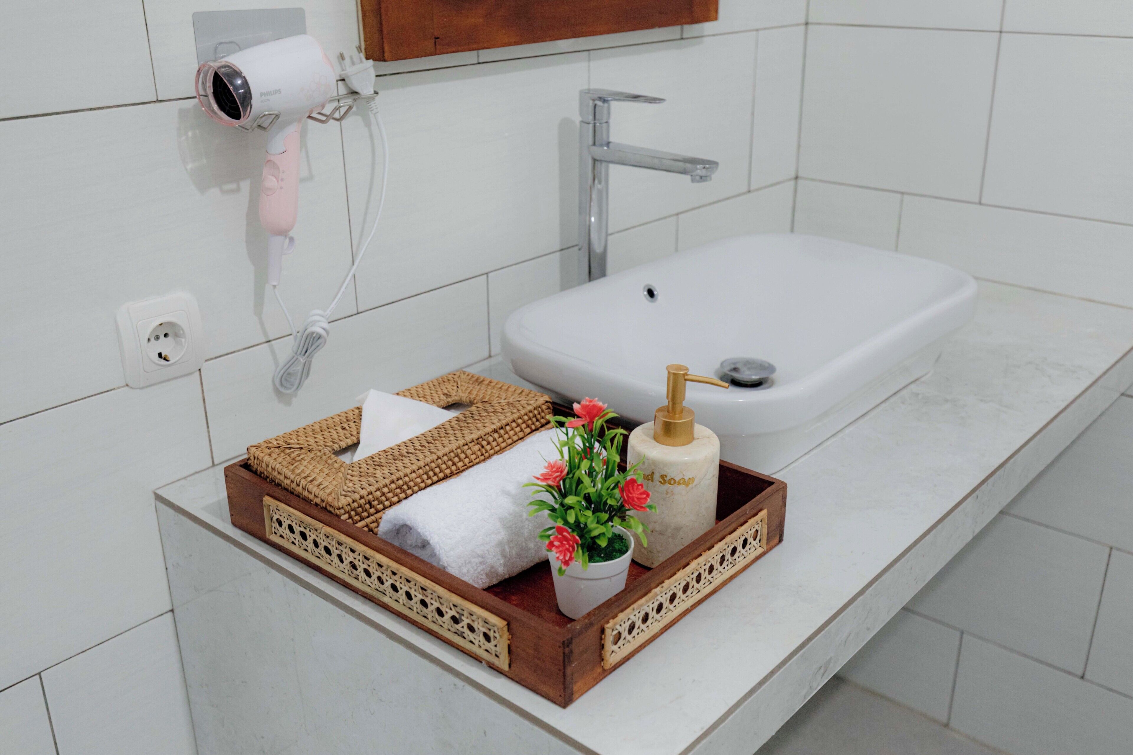 Ein Badezimmer mit einem weißen Waschbecken, einem Fön, einer Seifenfiole und einer Ablage mit Handtüchern und Blumen.