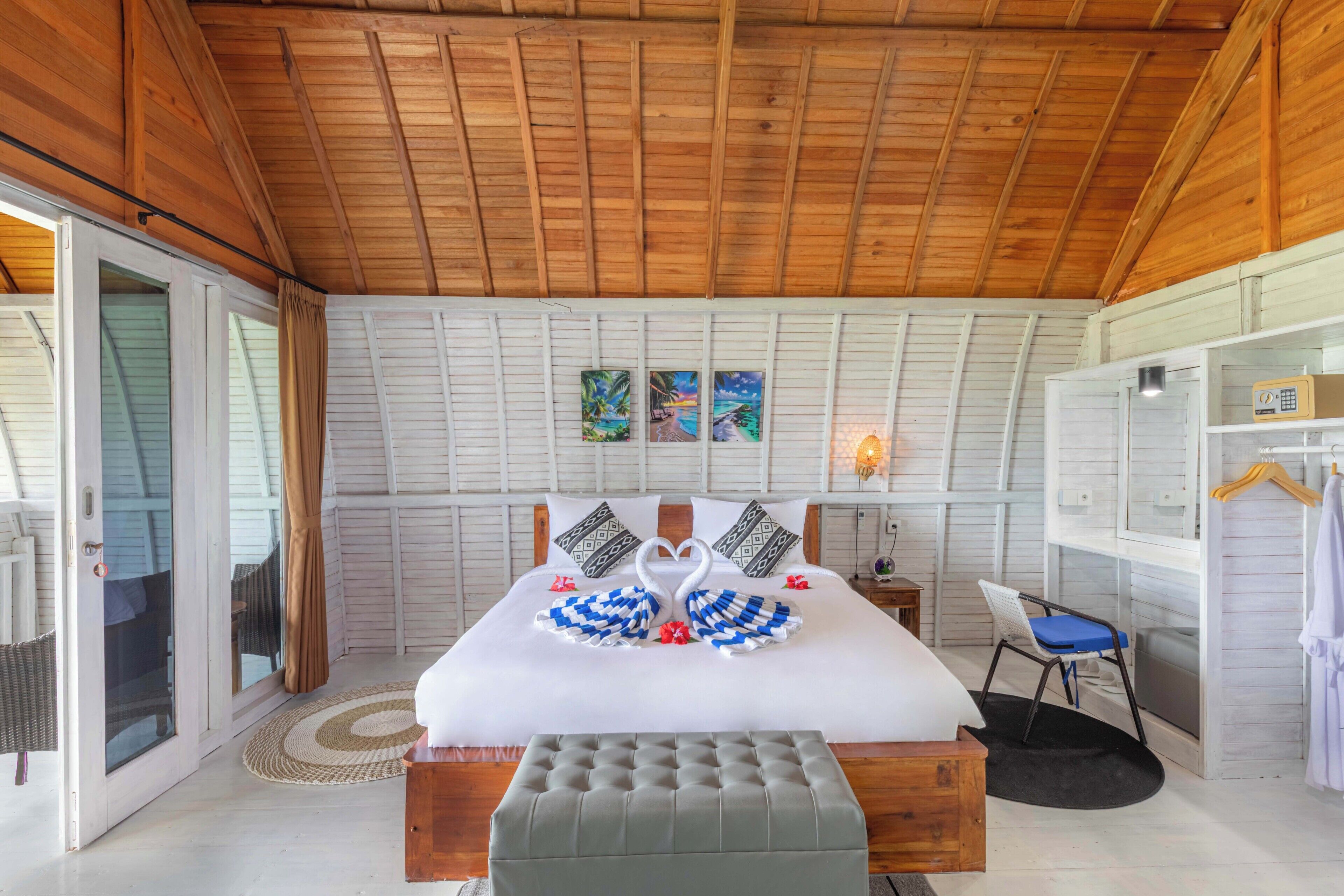 A bedroom with a wooden ceiling, a bed with a white bedspread, two pillows, a bench, a chair, and a small table.