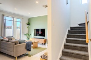 A modern living room with a sofa, TV, and staircase. - Brighton 3BR Townhouse |Sauna |Walk to Jetty,Train (Brighton)