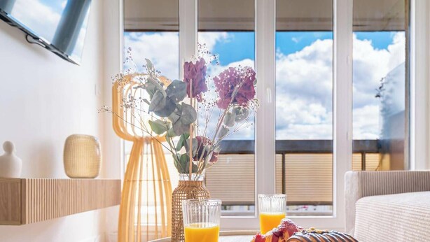 Ein Zimmer mit einem großen Fenster, einer Korbwase mit Blumen, einem Glas Orangensaft und einem Bett.