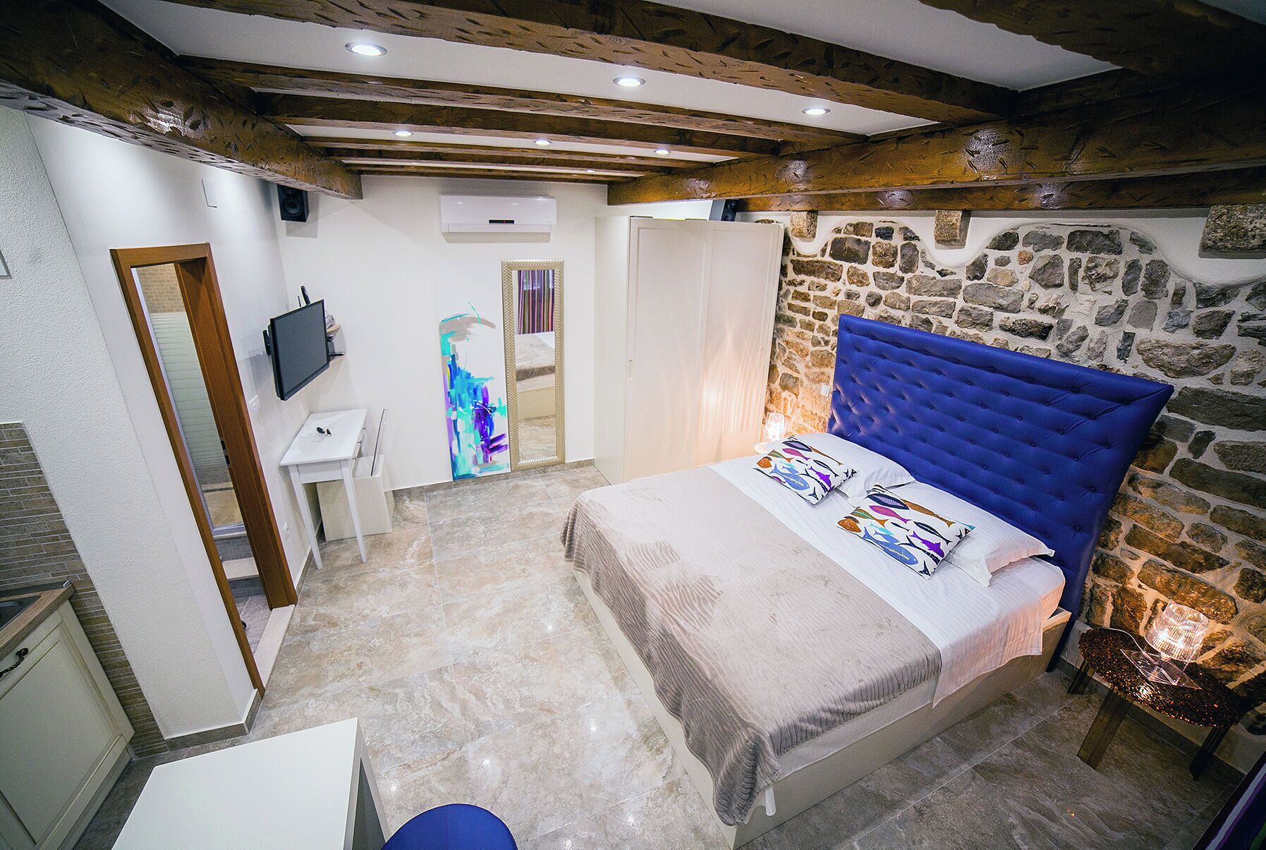 A bedroom with a stone wall, a bed with a blue headboard, a TV, and a small desk.