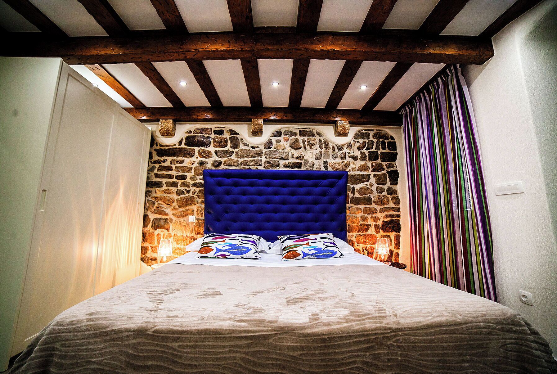 Un dormitorio con pared de piedra, cabecero azul y cortinas a rayas.
